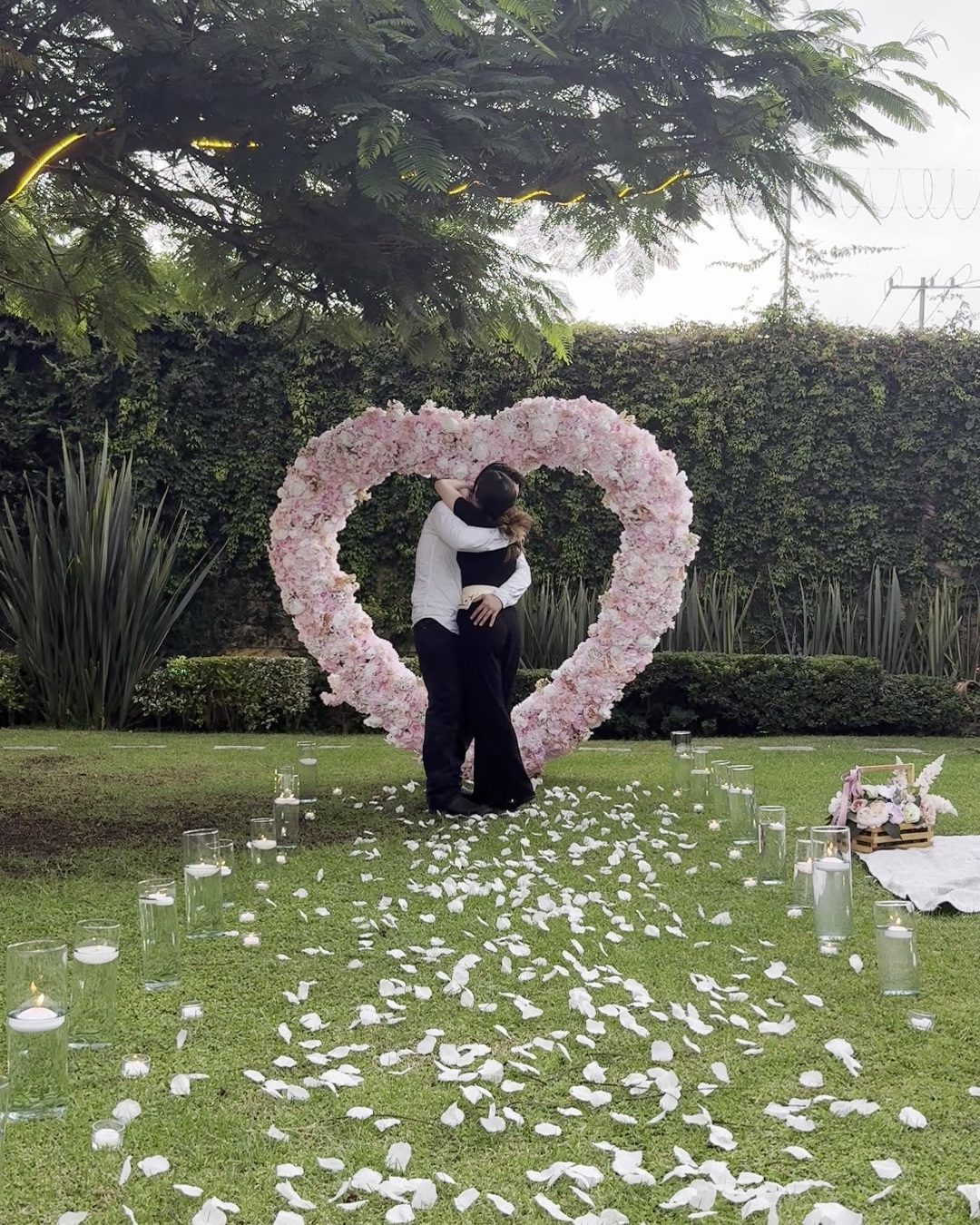 Pareja abrazándose dentro del corazón de flores, con el camino de pétalos y velas encendidas en el jardín